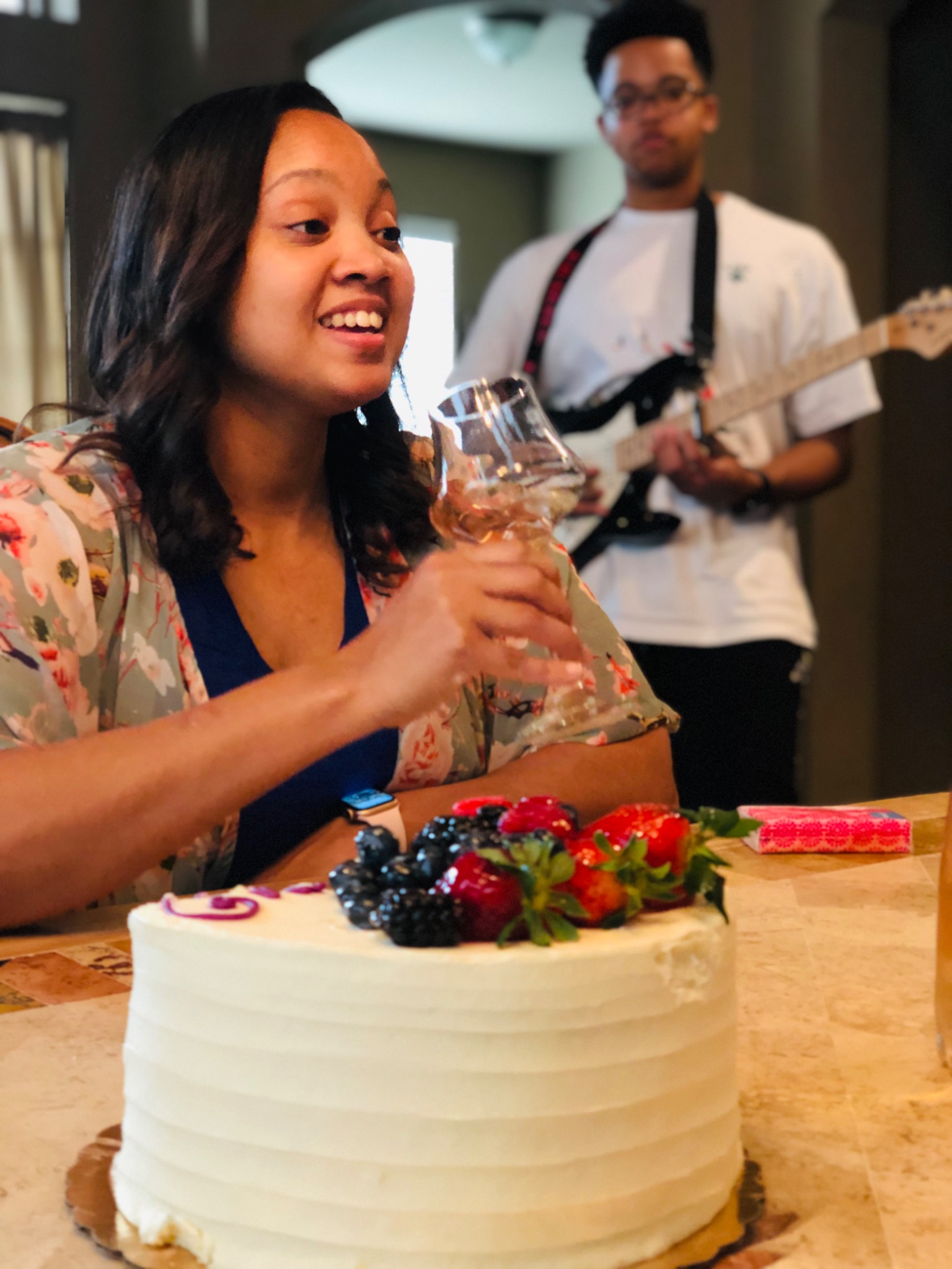 Christy smiling beside a birthday cake while her son plays guitar at home.